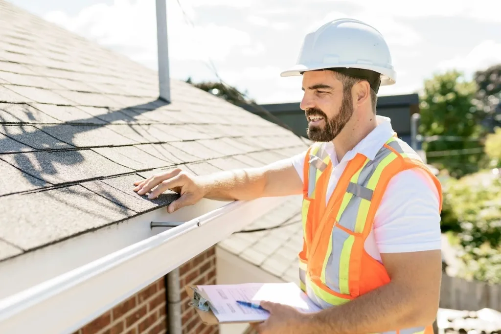 A professional roofer in safety gear inspecting the condition of shingles on a residential roof during a sunny day