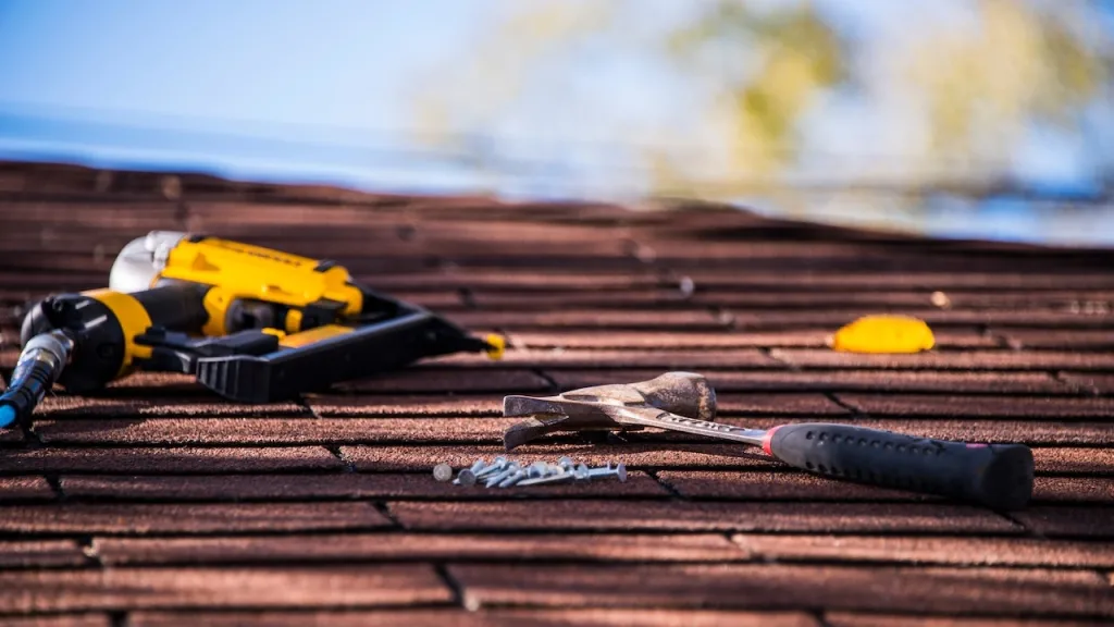 Roofing tools, including a hammer, drill, and nails, laid out on textured asphalt shingles, under a clear sky.