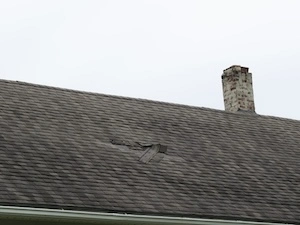 Roof with visible damage showing curled, cracked, and missing shingles, next to an old chimney, indicating severe wear and the need for immediate replacement.